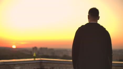 Back-view-a-male-freelance-programmer-in-a-hoodie-walks-on-the-roof-at-sunset-and-drinks-beer-looking-at-the-beautiful-view-of-the-city.